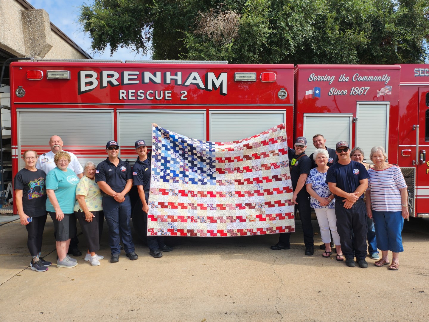group of quilters and fire fighters standing in front of a fire truck, holding up a quilt that looks like the American stars and stripes flag